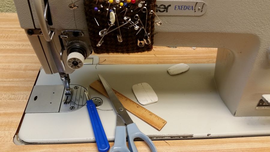 Tailor shop, sewing, alteration shop tools.  Seam Ripper, Bobbin, Push Pins, Safety Pins, Tailor Chalk, Ruler, and Scissors resting on a commercial sewing machine and sewing table. 
